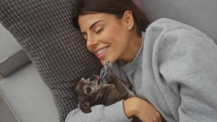 A smiling young woman cuddles with her chihuahua on a cozy couch inside a warm home interior.