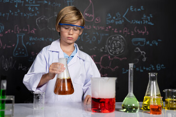 Blonde hair schoolboy in laboratory wear lab coat stand and learn science of chemistry technology in STEM class. The student hold beaker with orange liquid. On the table put many flask. Erudition.