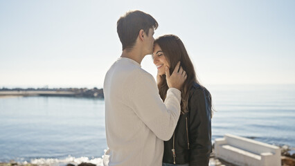 Fototapeta premium A man tenderly kisses a smiling woman's forehead at the sunny seaside, symbolizing love and affection between a couple.