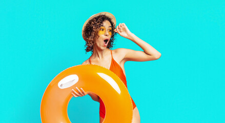 A young black woman wearing a straw hat, sunglasses, and an orange swimsuit holds a large orange inflatable ring in front of a bright blue background. She looks surprised and happy, copy space