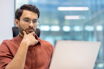 Businessman in glasses and casual attire working on laptop in modern office. Displays focused and...