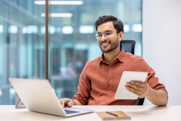 Young professional working at office desk using laptop and tablet, looks engaged and productive. Modern office environment with glass walls and natural light represents technology, business, and