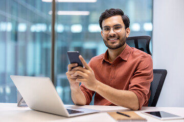 Confident businessman seated in modern office using smartphone and laptop, smiling. Professional atmosphere with open space and glass walls. Concept of business, technology, communication success.