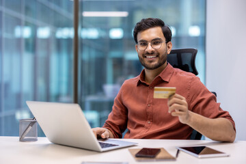 Business professional sitting at desk holding credit card using laptop in modern office. Engaged in...