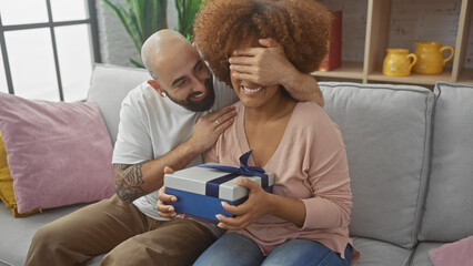 A man surprises a woman with a gift on a couch, sharing a joyful moment in a cozy home interior.