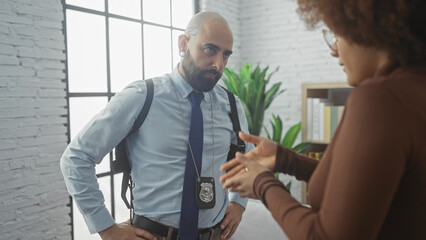 A bald man with a beard, wearing a blue shirt, tie, and police badge, talks to a woman inside a...