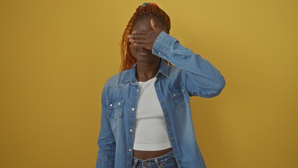 Young african american woman covering eyes with hand against a yellow background, evoking a 'see no evil' concept.