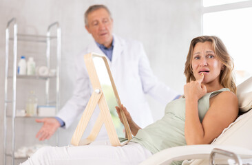 Obraz premium Upset female patient behaving discontentedly lying on medical chair with mirror in hands in aesthetic clinic