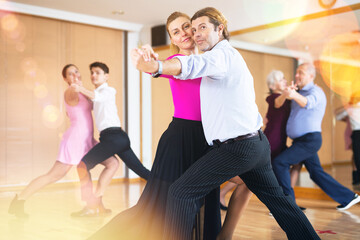 Obraz premium Positive adult couple enjoying slow foxtrot in dance studio. Amateur social dancing concept..