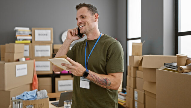 Smiling young man with a beard and tattoo talking on the phone while holding a notebook in a warehouse.