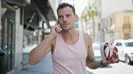 A young hispanic man in a pink tank top talks on a phone while holding a heart-shaped gift in an...