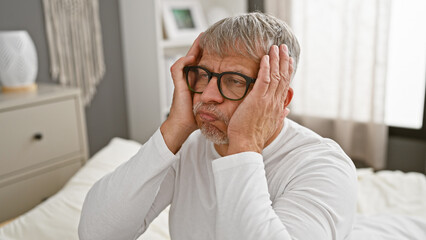 Mature grey-haired man in glasses looks pensive while sitting on a white bed in a well-lit bedroom,...