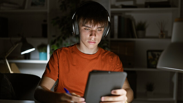Focused teen boy studying late at night in a home interior, using a tablet and headphones.