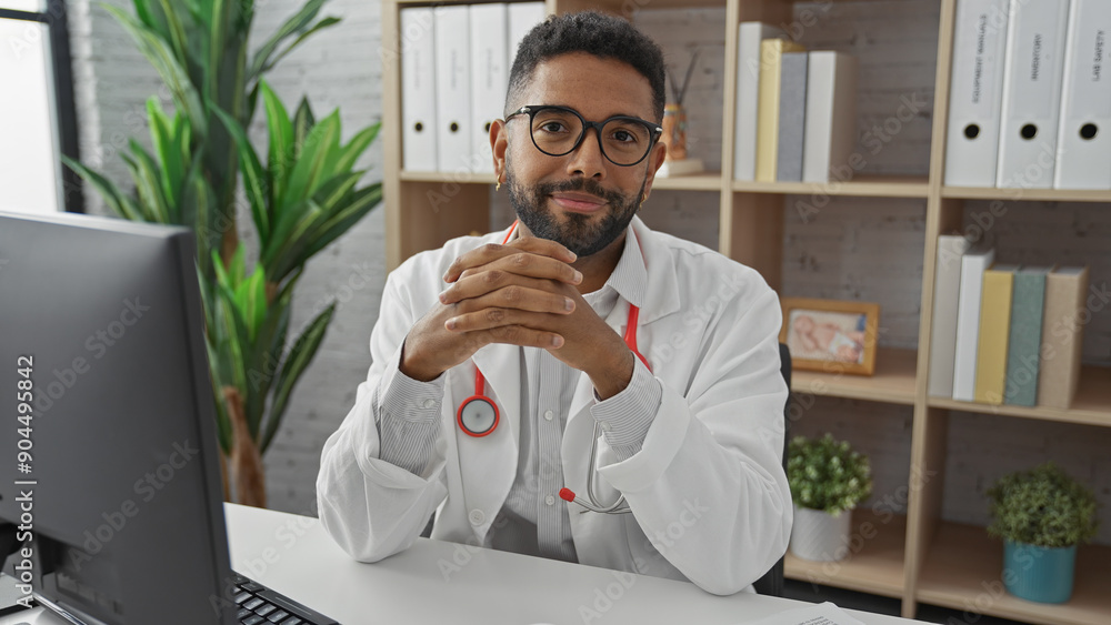 Wall mural A confident african american male doctor sits in a clinic office, dressed in a white coat with a stethoscope and glasses, working at a computer.