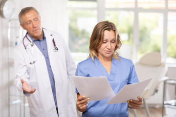 Upset shocked female medical worker in blue scrub standing in clinic office with papers in hands on...