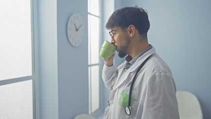 A young adult man with a beard wearing a lab coat drinks from a mug in a hospital corridor.