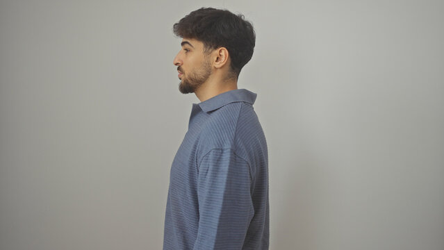 A young bearded man in a modern outfit poses sideways against a white background, exuding a casual and confident demeanor.