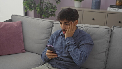 A young arab man with a beard looks at his phone in a modern apartment living room.