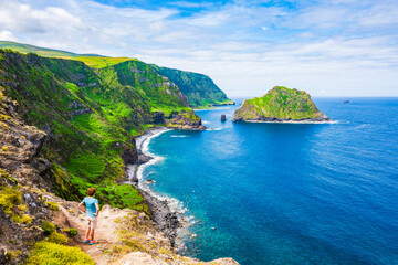 Young woman tourist looking at beautiful bay with beach and green cliffs at Baia de Alem, Flores...
