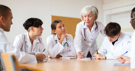 Fototapeta premium Experienced elderly female professor of medicine giving lecture to group of interested internship doctors sitting at same table in professional seminar..