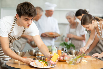 Attentive young male attendee of cooking course engaged in decoration of baked piece of salmon in the kitchen