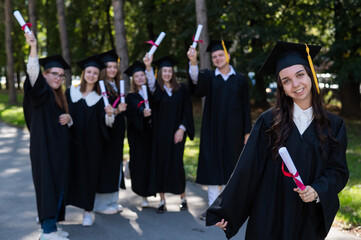 Group of happy students in graduation gowns outdoors. A young girl with a diploma in her hands in the foreground.