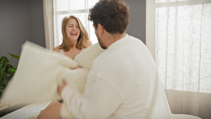 A joyful woman and man engage in a playful pillow fight in a sunlit bedroom, embodying love and happiness together in a domestic setting.