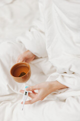 Person in bed with cup of tea and thermometer on bedside table