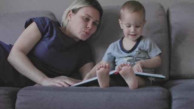Boy and mom sits comfortably on couch engaged in encyclopedia exploring new information. Curious boy on sofa and young mother reading encyclopedia discovering world around intellectual development