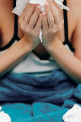 Sick woman blowing nose with tissue while sitting on bed with towel over head