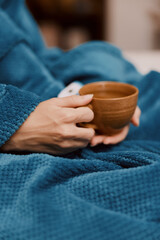 Person wrapped in blue blanket holding a cup of coffee and a remote control