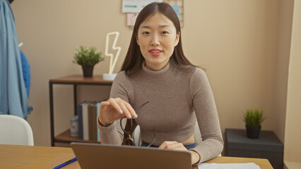 Naklejka premium A young asian woman with glasses working on laptop in a modern home office setting, conveys focus and professionalism.