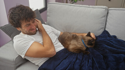 A young man in white shirt cuddles a siamese cat on a navy blue blanket on a sofa in a cozy room.