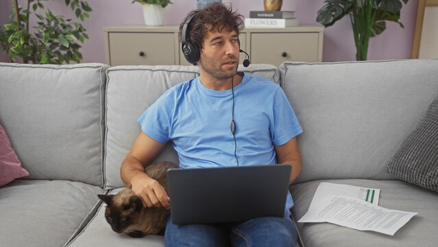Hispanic man with headphones using laptop and petting cat on sofa in a cozy living room. - Powered by Adobe