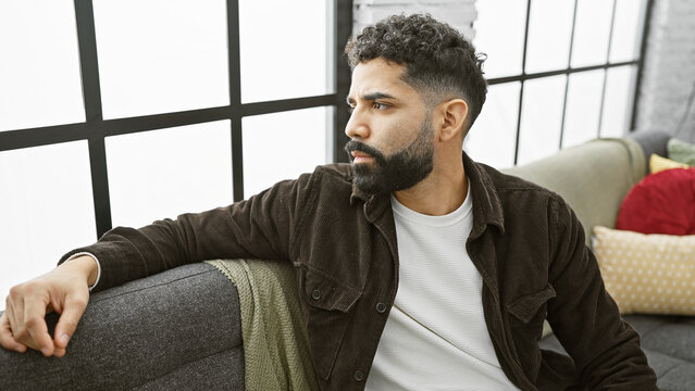 Handsome young man with beard sitting thoughtfully in a modern apartment living room.