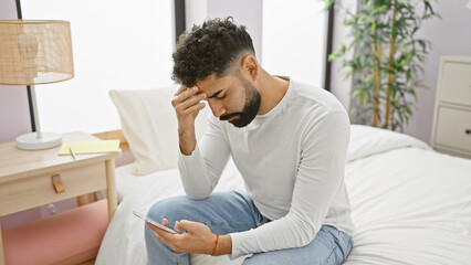 A worried young man with a beard sits on a bed at home, scrutinizing his smartphone in a modern bedroom