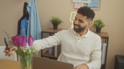 Fototapeta premium A cheerful young hispanic man takes a selfie in a modern home office with decor and laptop.