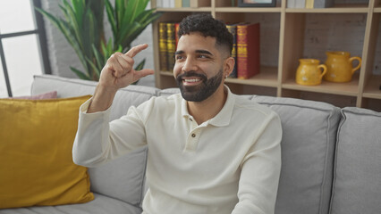 Smiling hispanic man gesturing small size indoors, on a neutral couch, in a cozy, modern living room.