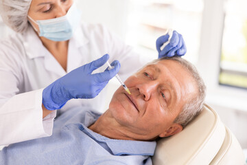Fototapeta premium Female doctor conducting face revitalization procedure for old man patient by micro-injection method