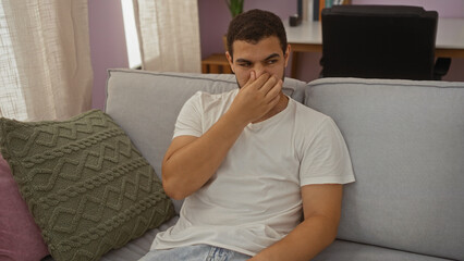 Young man sitting on a couch at home pinching his nose while looking uncomfortable in a cozy living...