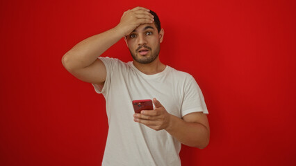 Young hispanic man holding a smartphone with a surprised expression over an isolated red background wall