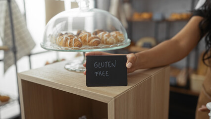 Woman displaying a gluten-free sign in a bakery with pastries under a glass dome, emphasizing the gluten-free options available.