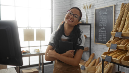 Asian woman with glasses and long hair holding a tablet, smiling in a bakery shop interior with...