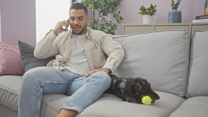 A relaxed hispanic man talks on the phone while sitting with his dachshund on a sofa in a cozy...