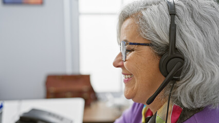 A smiling mature woman with grey hair wearing a headset indoors at her office workplace.