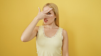 Young caucasian woman covers one eye with hand against a plain yellow background, showing playfulness and surprise.