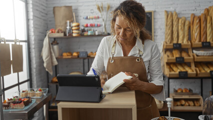 Middle-aged woman in a bakery writing in a notebook while checking a tablet surrounded by bread and pastries, wearing an apron, hispanic, indoors, focused on her task in a cozy shop environment.