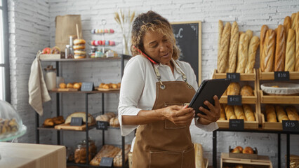 Woman working in a bakery talking on the phone and using a tablet surrounded by various breads and pastries