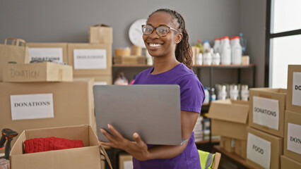 Smiling woman holding laptop in warehouse with donation boxes, portraying volunteer work and...