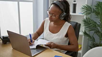 African american woman wearing headphones focuses on taking notes during her work in a modern indoor office.